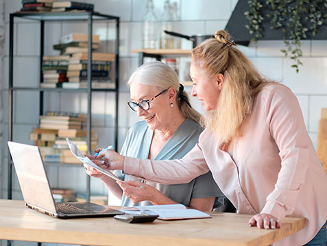 Senior woman using laptop for websurfing in her kitchen. middle-aged daughter helps her mother with documents. Mature lady sitting at work typing a notebook computer in an home office