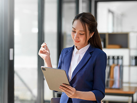 Attractive smiling Asian businesswoman standing holding tablet working and recording work details in office.