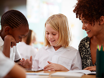 Female Teacher With Two Elementary School Pupils Wearing Uniform Using Digital Tablet At Desk