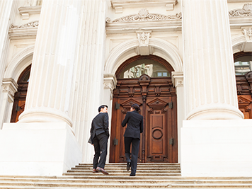 Two people dressed in black suits walking up the stairs into a government building